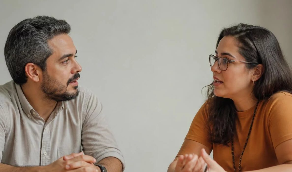 Três participantes conversam ao redor de uma mesa comprida numa sala clara de janelas altas